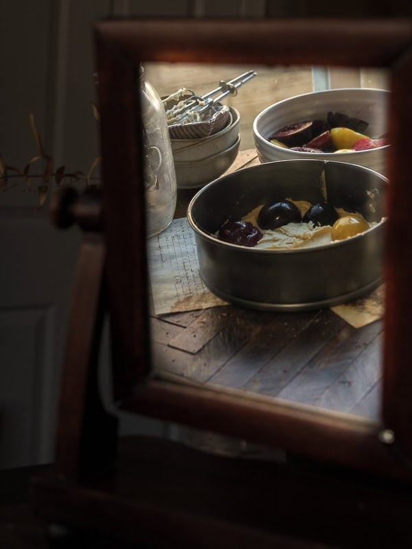 A wooden-framed mirror reflects two metal baking pans filled with colorful ingredients, sitting on a rustic wooden table with a vintage egg beater and glass jar in the background.